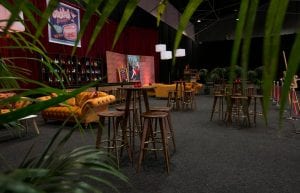 cocktail bar with posters on faux brick wall. orange velvet sofas and wooden hi bars and stools in front. red drape walls and copper pressed tin bar