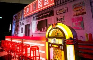 12m diner scene backdrop. red and white illuminated bar counter in front with red plastic ghost stools. black and white chequered dance floor and wurlitzer jukebox