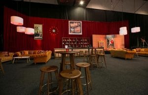 cocktail bar with posters on faux brick wall. orange velvet sofas and wooden hi bars and stools in front. red drape walls and copper pressed tin bar