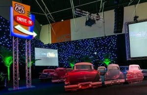 star cloth wall and palm trees. drive in cinema set inside a ballroom. car cutouts with sofas in front and drive in speakers for guests to watch movies