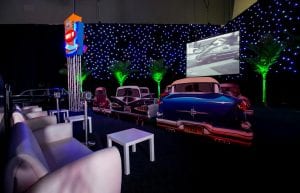 star cloth wall and palm trees. drive in cinema set inside a ballroom. car cutouts with sofas in front and drive in speakers for guests to watch movies