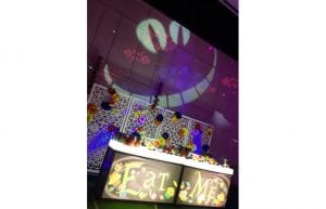 eat me print on the front of an illuminated bar for a dessert buffet. lighting gobo shape of cheshire cat on wall. alice in wonderland theme