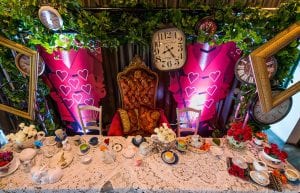 mad hatter tea party table styling with lace table cloths and teapots, cups and saucers. card soldier flat props and greenery with hanging clocks and frames