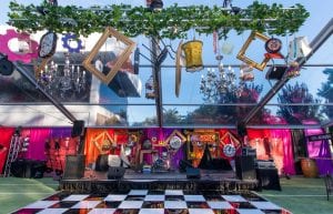 alice in wonderland themed event in clear roof marquee with ceiling piece made up of hanging clocks, gold frames, lampshades and black and white chequered dance floor. stage backdrop with coloured silk drapes