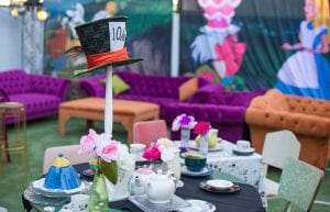 pink and orange velvet sofas and ottomans. table set up as mad hatters tea party with lace tablecloths, teacups and pots, saucers and flowers in vases. alice in wonderland theme