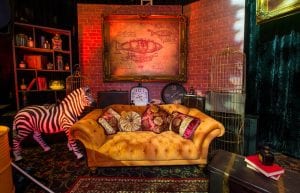 Steam Punk event theme with large gold frames, bookcase, zebra statue, bird cages, clocks, trunk, suitcases, books, styling props, orange velvet chesterfield sofa, Arabian rug, cushions, black drapes, brick wall props, trussing and steam punk prints for hire.